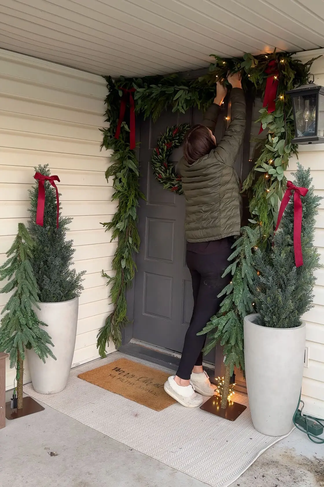 A woman adding twinkling string lights to greenery garland on a holiday-decorated front porch, featuring festive red bows, a greenery wreath on a gray door, potted evergreens, and small lit Christmas trees, creating a warm and inviting glow.